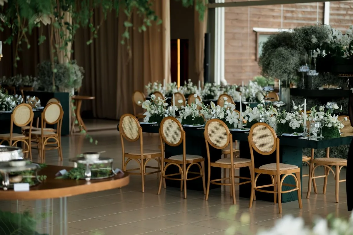 Mesa de evento decorada com flores brancas e velas, cadeiras de vime e bandejas de buffet ao fundo