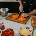 Chef cortando cenoura em tábua de madeira, cercado de tomate, cebola, alho e carne em cozinha profissional