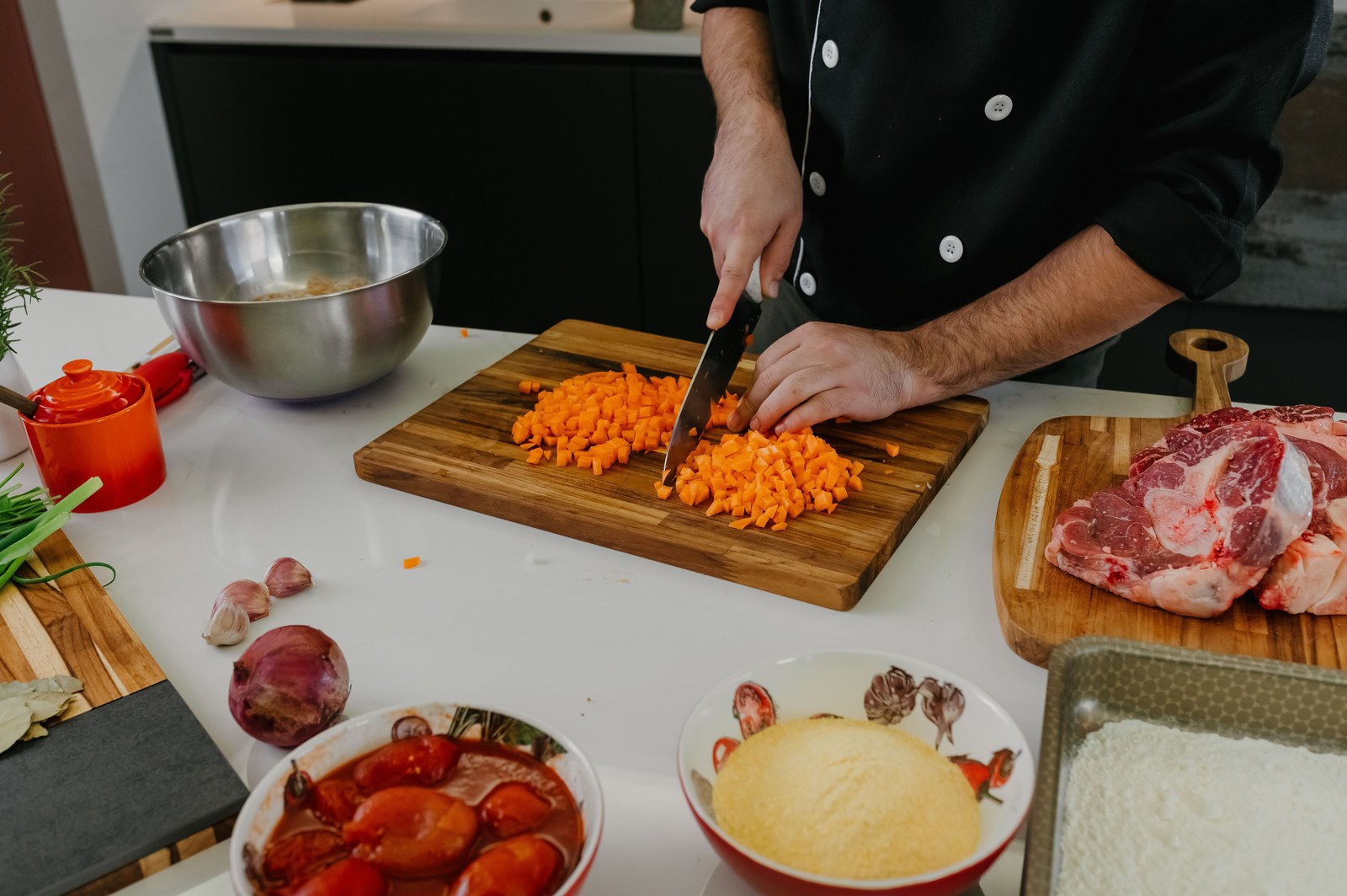 Chef cortando cenoura em tábua de madeira, cercado de tomate, cebola, alho e carne em cozinha profissional