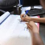Pessoa anotando um compromisso com caneta azul num calendário de mesa, ao lado do teclado do computador