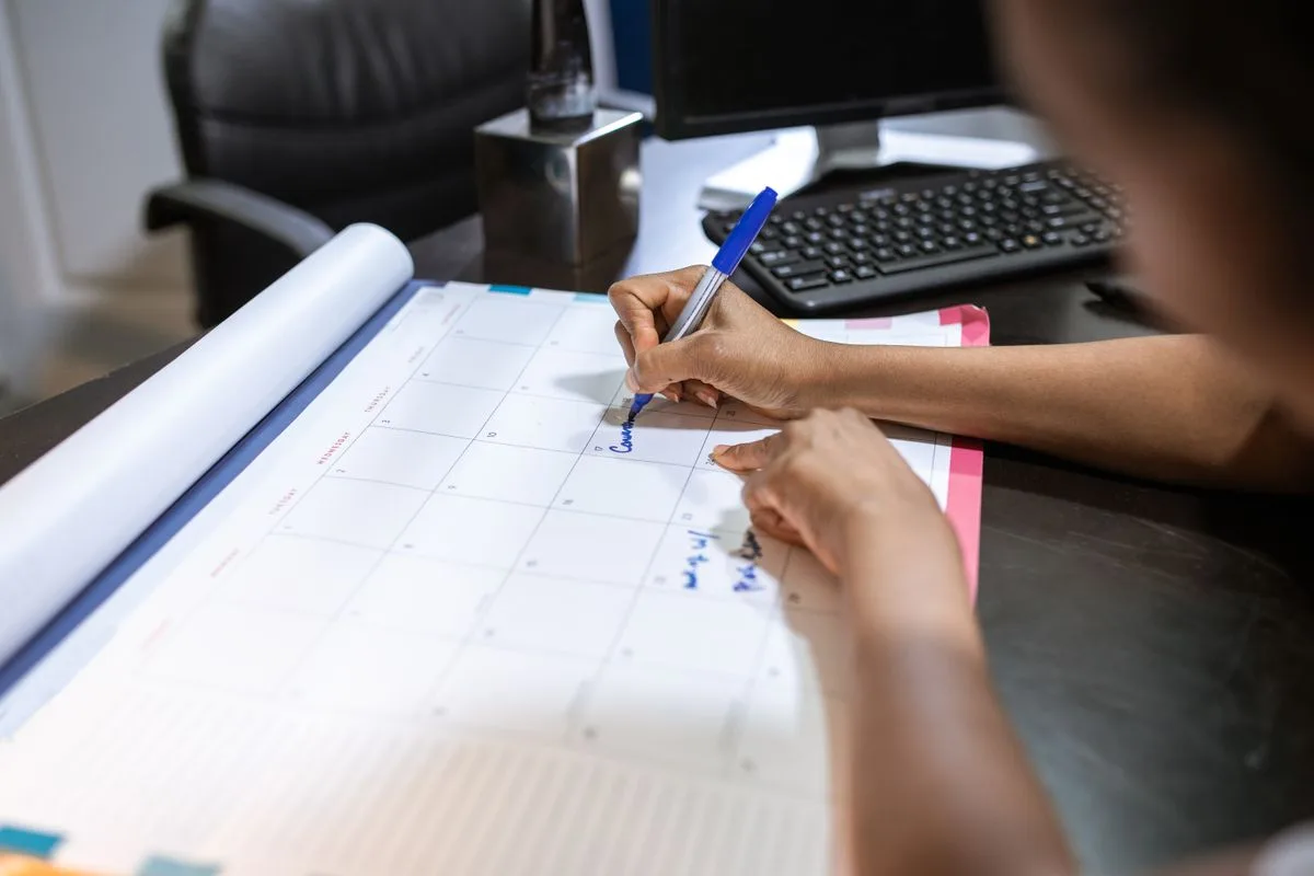 Pessoa anotando um compromisso com caneta azul num calendário de mesa, ao lado do teclado do computador